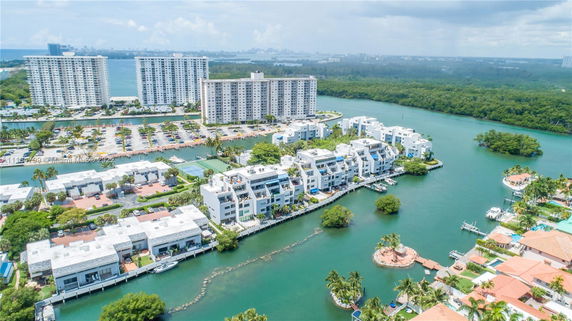 Aerial view of multiple residential buildings alongside a waterway, surrounded by greenery and distant skyscrapers.