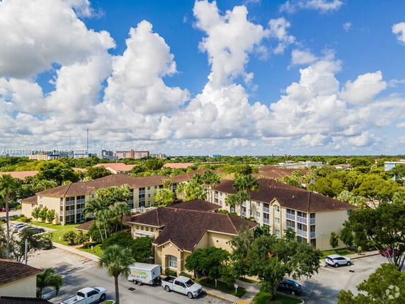 Aerial view of a residential complex with multiple multi-story buildings, surrounded by trees and greenery under a blue sky with clouds.