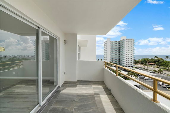 Balcony view from an apartment with a glass sliding door, showing nearby high-rise buildings and a view of the ocean.