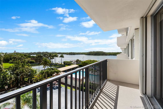 Balcony view with railing overlooking trees, a tennis court, and water under a blue sky.