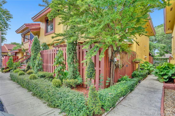 Front view of a two-story house with yellow walls and red roofs, surrounded by greenery and a wooden fence.