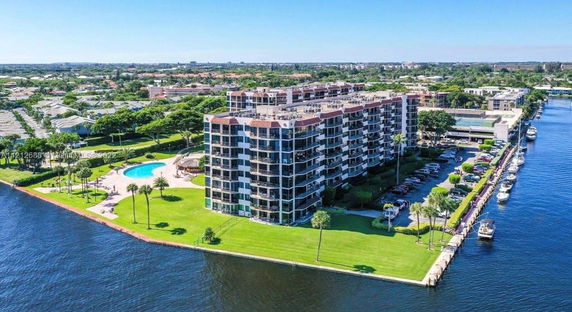 Aerial view of a multi-story residential building complex with a pool, situated by a waterfront. The complex is surrounded by green lawns and parking areas.