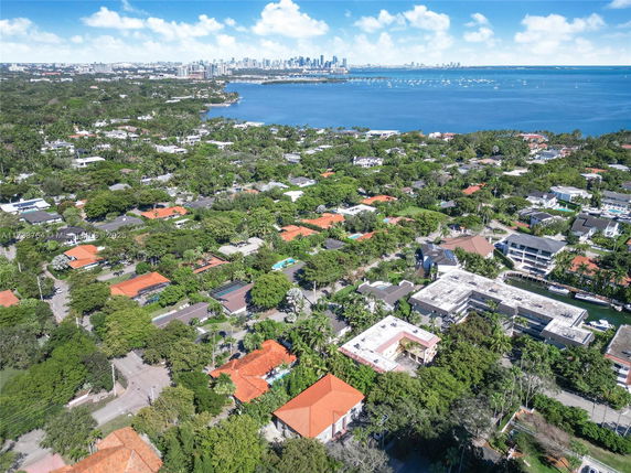 Aerial view of residential area with numerous houses and buildings surrounded by greenery, overlooking a body of water with a city skyline in the distance.