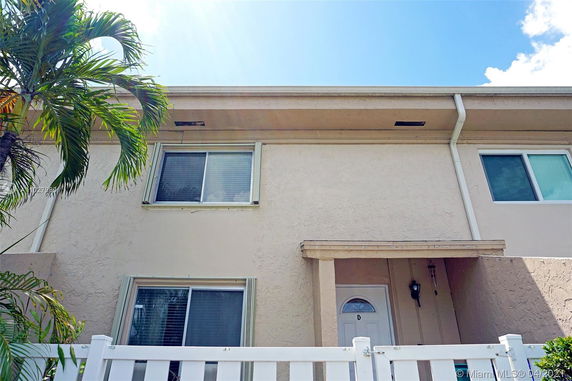 Front view of a two-story house with a light exterior and white fence, featuring multiple windows and a small entrance.