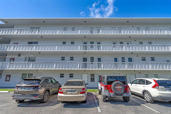 Front view of a multi-story apartment building with balconies and cars parked in front.