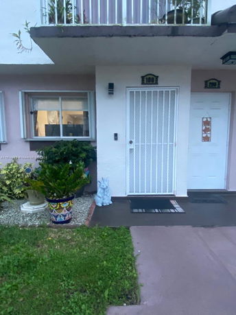 Front view of a residential unit with a white door and barred security screen, a window, and potted plants outside.