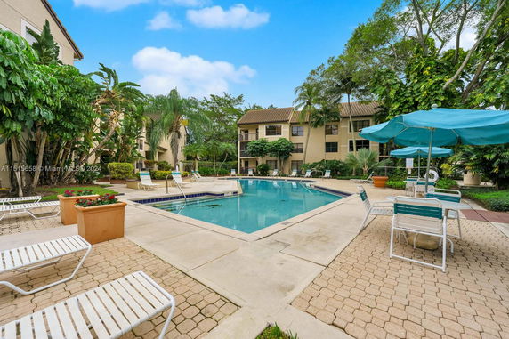 Outdoor swimming pool area with surrounding lounge chairs and umbrellas, adjacent to residential buildings.
