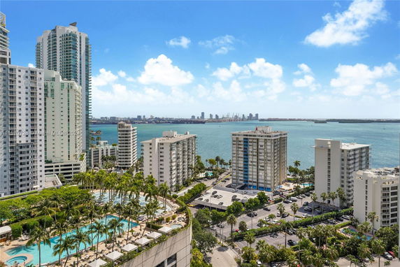 Aerial view of a cityscape with tall buildings near a waterfront and a pool area surrounded by palm trees.