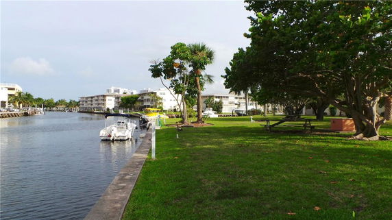 View of waterfront area with boats, grassy area, trees, and surrounding buildings.