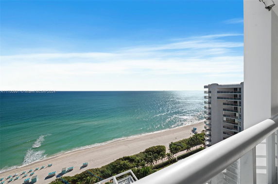 View from a balcony overlooking the ocean and beach, with a high-rise building visible.