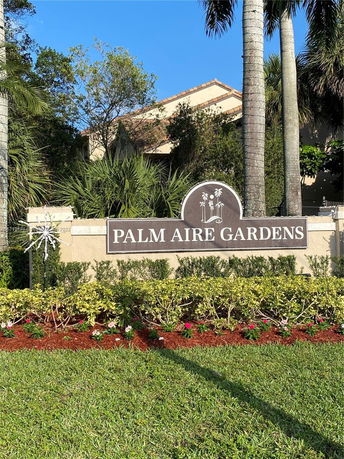Front view of Palm Aire Gardens entrance sign with a building partially visible behind trees.