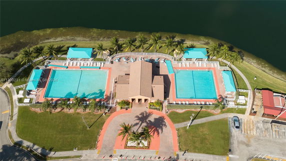 Aerial view of two large outdoor swimming pools and a central building surrounded by poolside seating and palm trees.