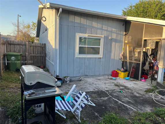 Side view of a small building with a fenced area and outdoor patio. There is a barbecue grill and a striped chair on the patio.