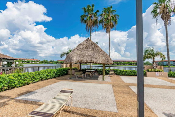 Outdoor seating area with a thatched roof structure and palm trees, overlooking a water body.