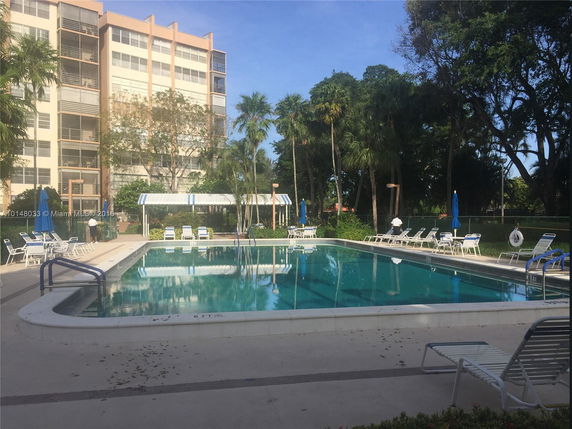View of an apartment building adjacent to a swimming pool surrounded by lounge chairs and palm trees.