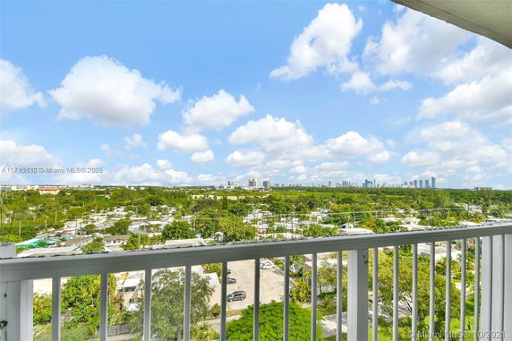 View from a balcony overlooking a residential area with buildings and greenery, under a sunny sky with clouds.