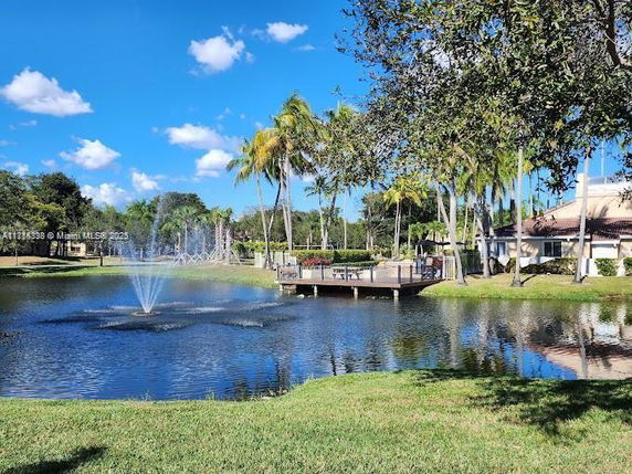 View of a pond with a fountain and surrounding trees and buildings.
