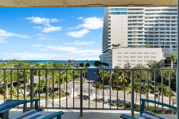 View from a balcony overlooking a street with palm trees and a large modern building in the background, with the ocean visible beyond.