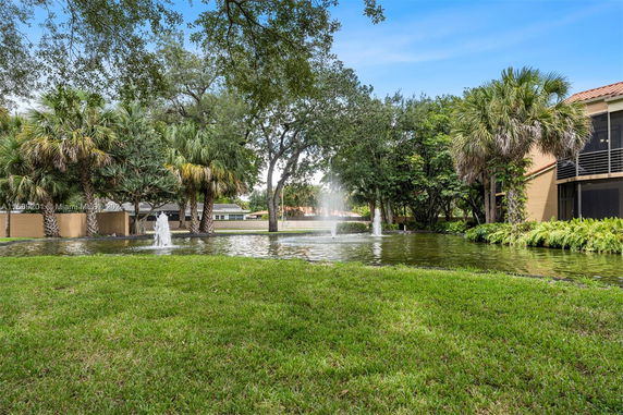 View of a landscaped area with a pond and fountains in front of buildings.