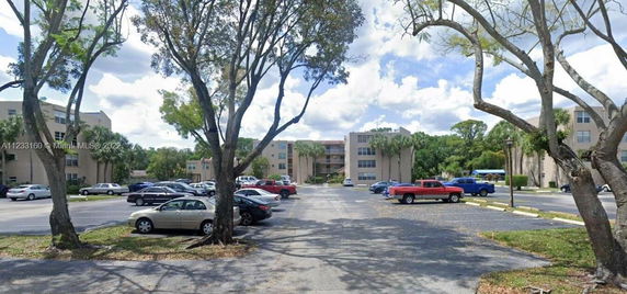 Outdoor parking area in front of a multi-story building complex with parked vehicles and trees.