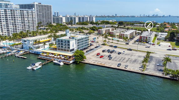 Aerial view of a waterfront building with a marina and parking area, surrounded by other buildings and a body of water.