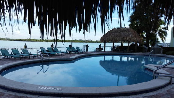 View of a swimming pool with lounge chairs and a tiki hut, overlooking a waterfront area.