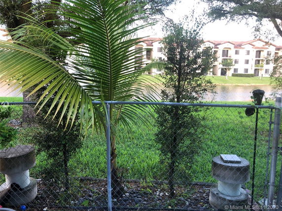 View of surrounding grassy area with trees and a chain-link fence, overlooking water in front of four-story buildings.