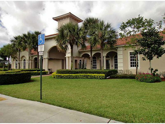 Front view of a building with arched windows and entrance, surrounded by palm trees and landscaped hedges.