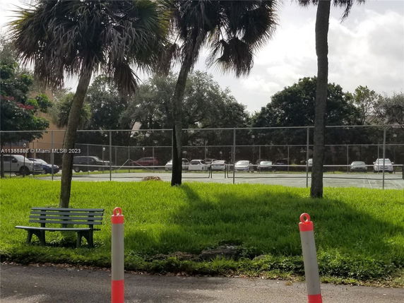 View of a fenced tennis court with parked cars in the background, surrounded by trees and grass.