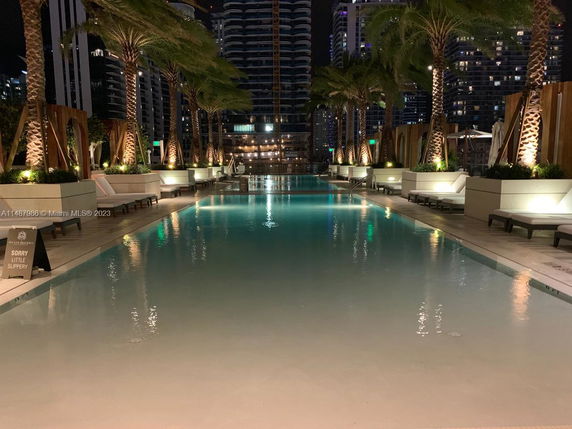 Rooftop with a lit swimming pool surrounded by lounge chairs and palm trees at night.