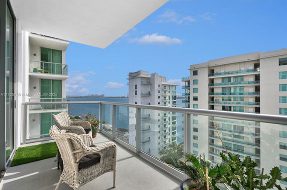 A balcony with wicker chairs and glass railing overlooking high-rise buildings and a body of water.