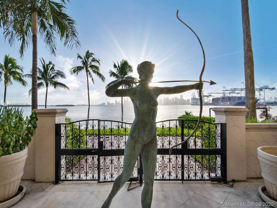 A waterfront view with a decorative statue holding a bow and arrow, overlooking the ocean with distant city skyline and cargo cranes.