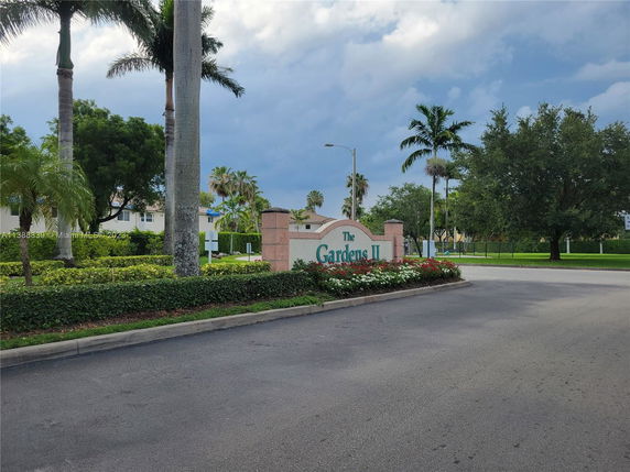 Entrance area with sign for 'The Gardens II', surrounded by palm trees and greenery.