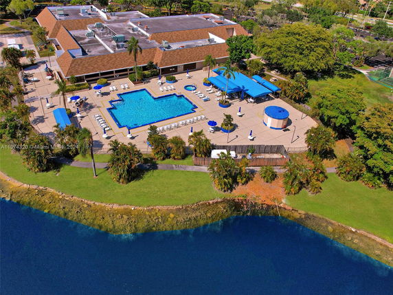 Aerial view of a building with a large swimming pool and lounging area, surrounded by greenery and water.