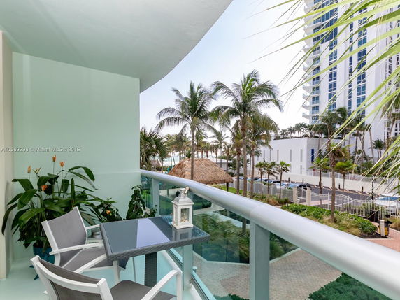 Balcony with outdoor seating overlooking palm trees and a tall building.