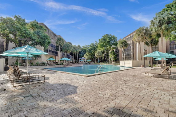 Outdoor swimming pool area with lounge chairs and umbrellas surrounded by buildings and trees.