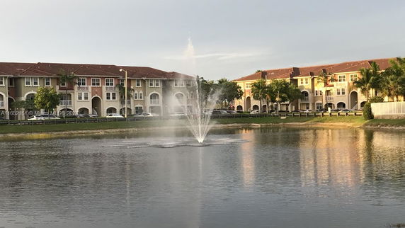 View of a multi-story residential complex with a red roof, seen from across a pond with a fountain.