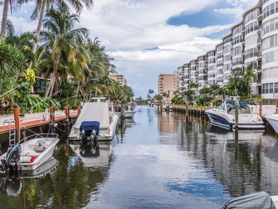 View of a canal lined with boats and palm trees, adjacent to multi-story residential buildings.