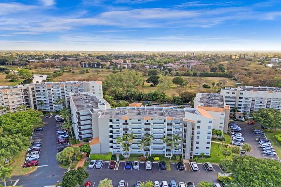 Aerial view of a multi-story residential complex with nearby parking and trees. The image captures the complex's rooftops with surrounding residential and green areas in the distance.