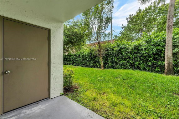View from a porch area with a closed door, overlooking a grassy yard surrounded by trees and bushes.