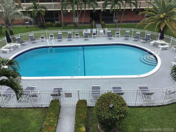 View of a swimming pool surrounded by lounge chairs and greenery, adjacent to a building.