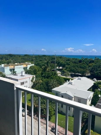 Panoramic view from a building balcony overlooking other buildings, trees, and the sea in the distance.
