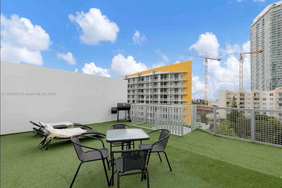 Rooftop area with artificial grass, outdoor furniture, and a barbecue grill overlooking nearby buildings and construction cranes.