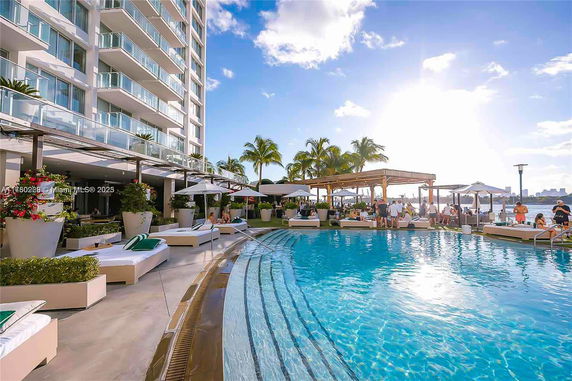 Outdoor swimming pool area with lounge chairs and umbrellas next to a multi-story building.