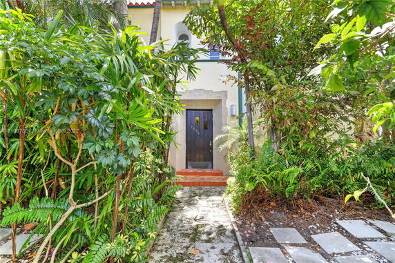 Front view of a house with a stone-framed door and a pathway surrounded by lush greenery.