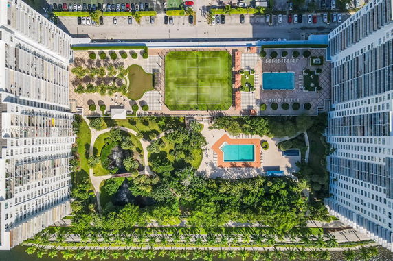 Aerial view of a recreational outdoor area with a tennis court, two swimming pools, garden paths, and landscaped greenery between two buildings.