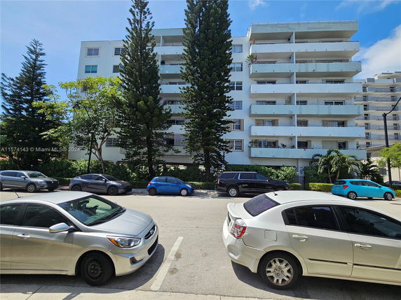 Front view of a multi-story apartment building with balconies and surrounding tall trees.