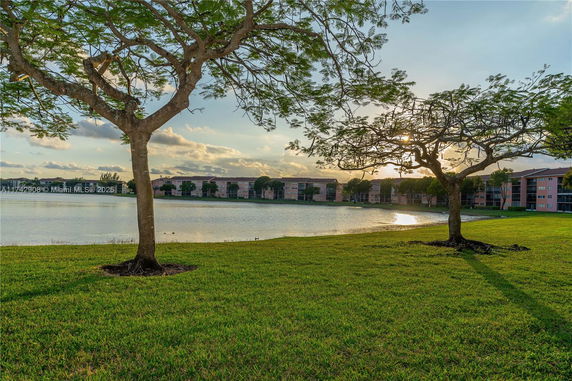 View of buildings across a lake with trees and grassy area in the foreground.