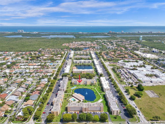 Aerial panoramic view of a residential area with multiple apartment buildings, a small lake, and surrounding greenery leading to a distant ocean.