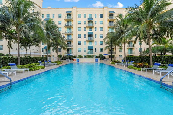 Outdoor swimming pool with lounge chairs and palm trees in front of a multi-story building.